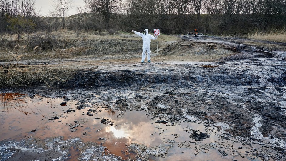 Человек в спецкостюме стоит рядом с почвой и водой, качество которых ухудшилось из-за выхлопных газов Человек в спецкостюме стоит рядом с почвой и водой, качество которых ухудшилось из-за выхлопных газов