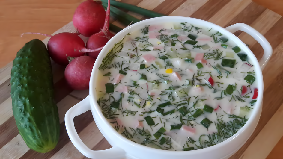 Супница окрошки стоит на столе рядом с огурцами и редисом Супница окрошки стоит на столе рядом с огурцами и редисом