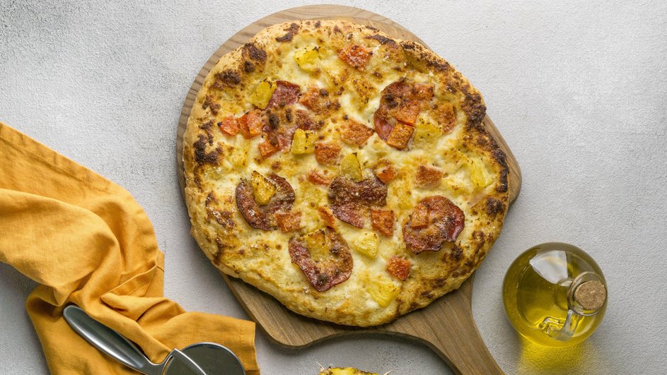 Гавайская пицца с ананасами и курицей лежит на доске рядом с ананасом и растительным маслом