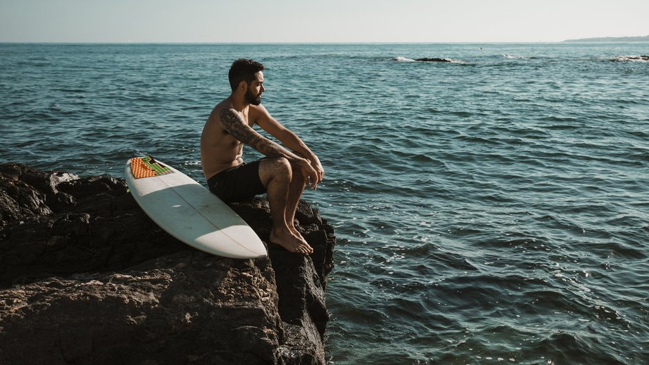 Мужчина сидит на камне над водой, рядом лежит доска для серфинга