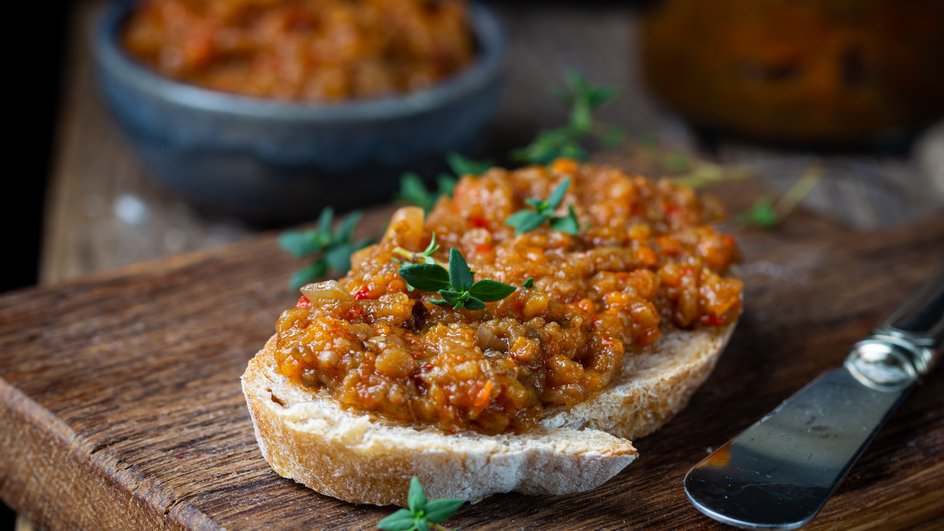 Икра из баклажанов с зеленью, поданная на кусочке хлеба, лежит на деревянной доске рядом со столовым ножом, тарелочкой и банкой икры 
