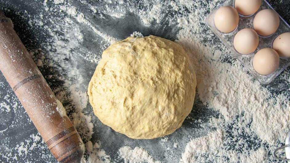 Заварное тесто лежит на столе с мукой рядом с яйцами и скалкой