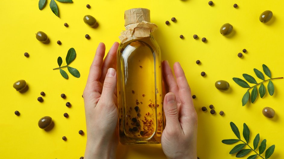 Бутылка оливкового масла, которую держат две руки, лежит на желтом фоне в окружении оливок, горошин черного перца и зеленых листьев Бутылка оливкового масла, которую держат две руки, лежит на желтом фоне в окружении оливок, горошин черного перца и зеленых листьев