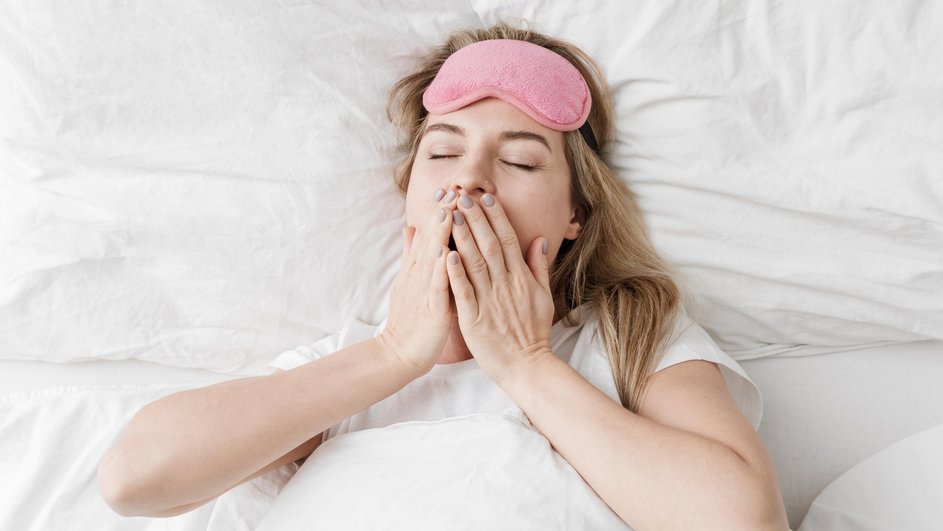 Женщина на белом постельном белье в розовой маске для сна, поднятой на лоб, но с закрытыми глазами, приложила руки к губам.