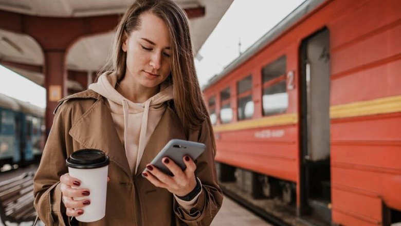 Женщина с кофе в руках ищет билет для путешествие на поезде в телефоне.