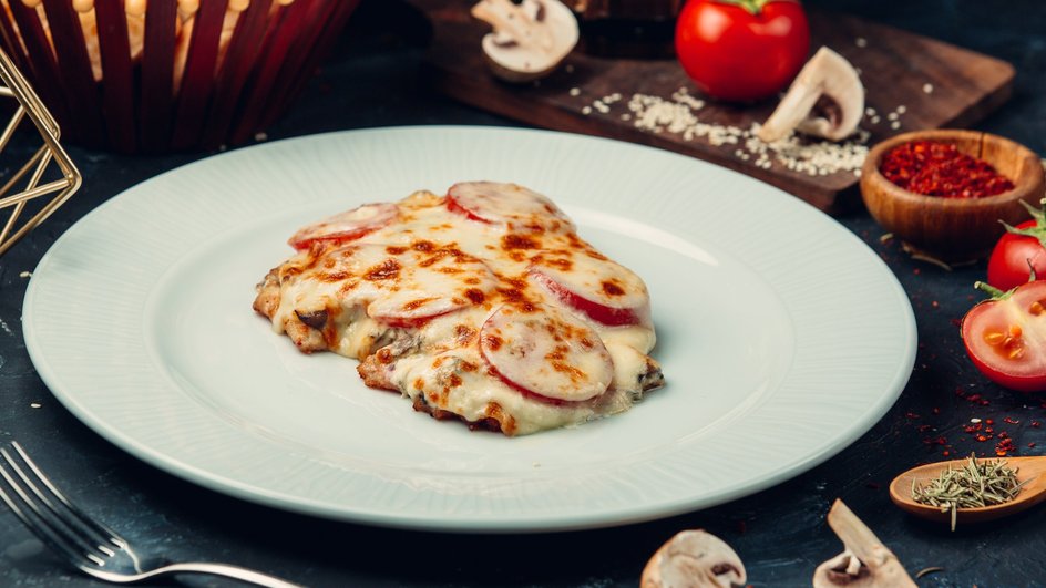 Мясо по-французски с курицей, грибами и томатами лежит на тарелке рядом с овощами