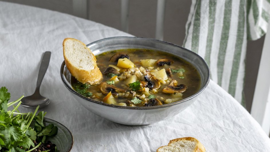 Вкусный суп из шампиньонов и мяса в мультиварке в серой тарелке стоит на столе рядом с зеленью, хлебом и ложкой