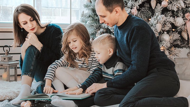 Семья с двумя детьми листает книгу на фоне новогодней ели
