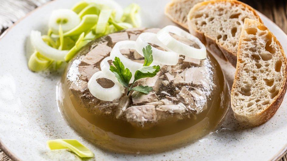Холодец из говядины, приготовленный в сковородке и украшенный кольцами лука и зеленью, лежит на тарелке рядом с кусочками хлеба Холодец из говядины, приготовленный в сковородке и украшенный кольцами лука и зеленью, лежит на тарелке рядом с кусочками хлеба