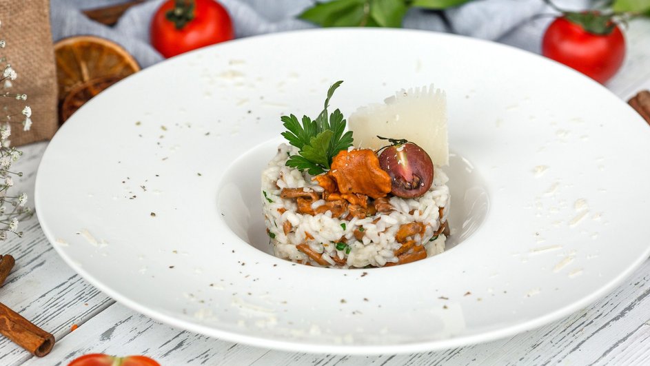 Ризотто с лисичками, украшенное листом петрушки, кусочком сыра и половинкой помидора, поданное в глубокой белой тарелке