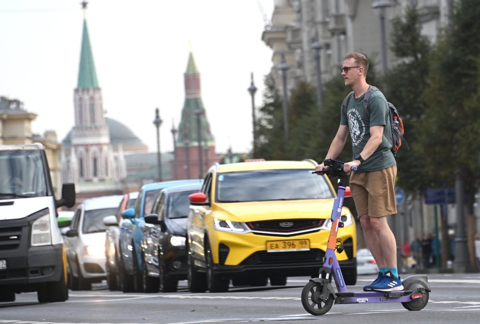 Мужчина пересекает дорогу на электросамокате в Москве. На заднем плане стоит автомобиль такси