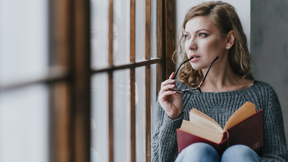 Девушка-Телец держит очки и книгу и смотрит в окно Девушка-Телец держит очки и книгу и смотрит в окно