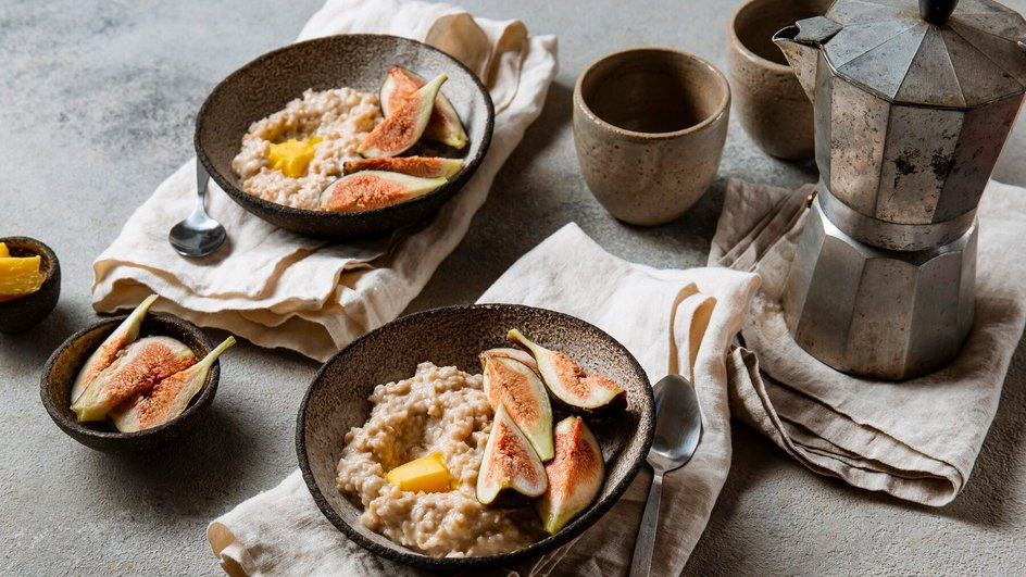  Две тарелки с овсяной кашей, украшенные инжиром и кусочками манго, на столе рядом с кофейником и чашками.