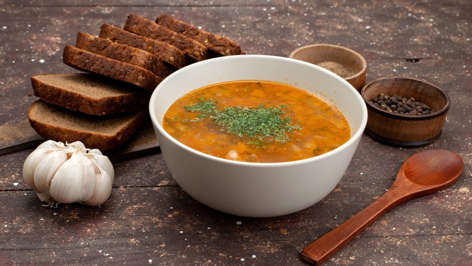 Тарелка гречневого супа с мясом стоит на столе рядом с кусочками хлеба, головкой чеснока и специями