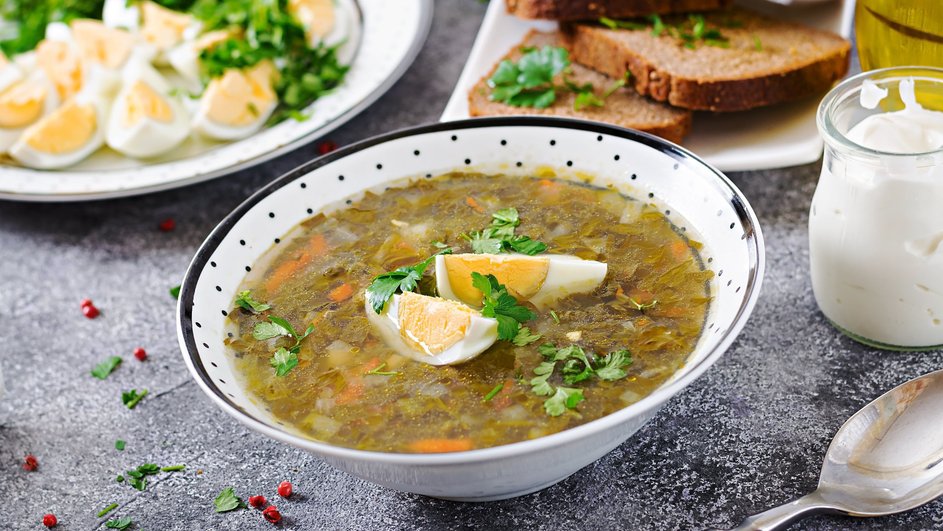 Зеленые щи с яйцом, украшенные зеленью, в глубокой тарелке стоят рядом с банкой сметаны, столовой ложкой, тарелкой с нарезанными отварными яйцами, зеленью и тарелкой нарезанного черного хлеба на столе