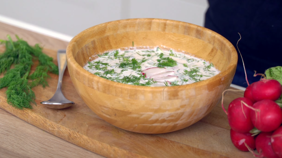 Тарелка окрошки на сметане с курицей и майонезом стоит на столе рядом с зеленью и редисом