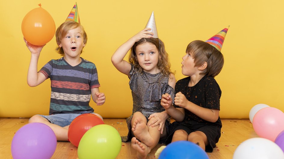 Трое детей - два мальчика и девочка - сидят на полу в праздничных колпачках, один из мальчиков держит в руке оранжевый шарик, на полу лежат другие разноцветные шары.