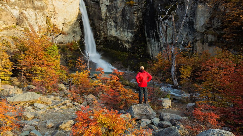 Человек в красной куртке стоит в горах на фоне водопада