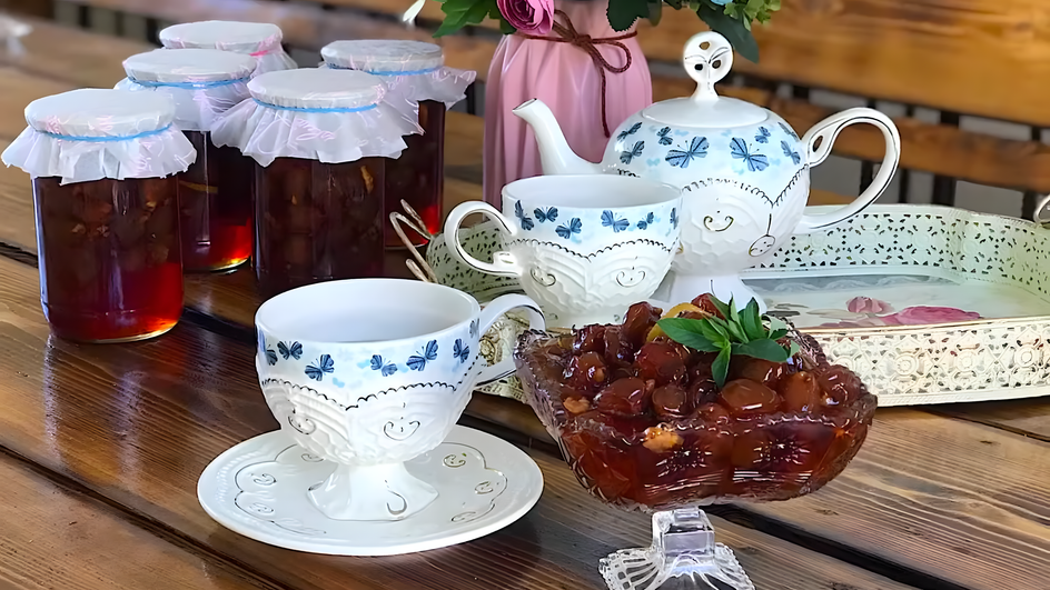 Креманка и банки варенья из белой черешни с грецким орехом стоят на столе рядом с чашками чая Креманка и банки варенья из белой черешни с грецким орехом стоят на столе рядом с чашками чая