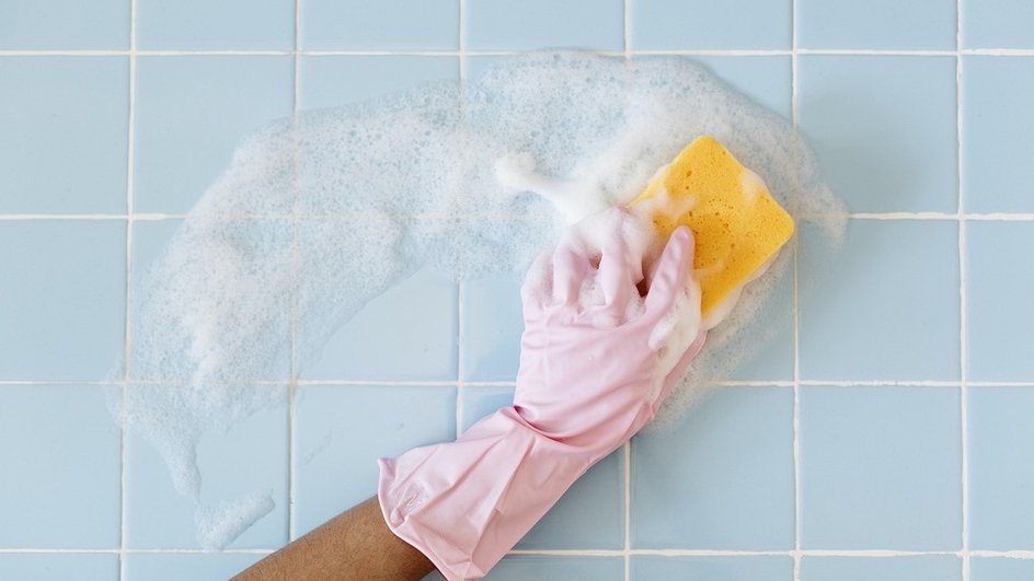 Рука в розовой перчатке желтой губкой моет плитку в ванной