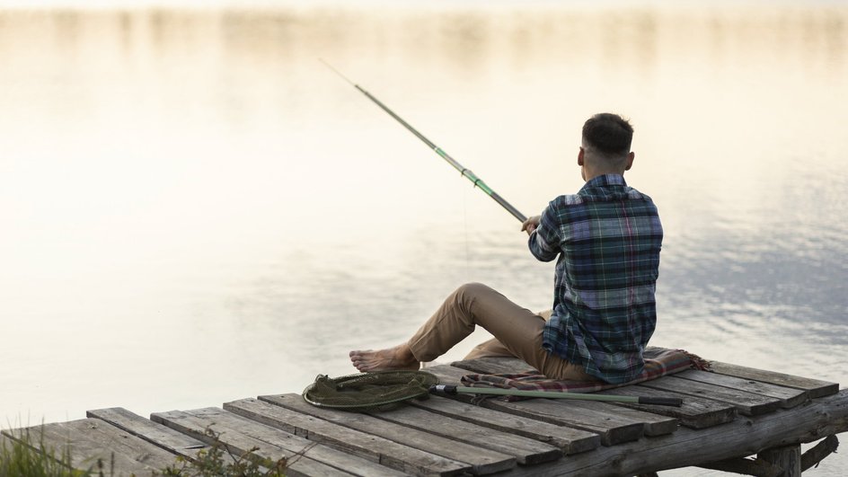 Мужчина держит удочку сидя на пирсе на фоне реки