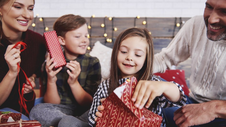 Семья с двумя детьми открывает подарки на фоне гирлянды