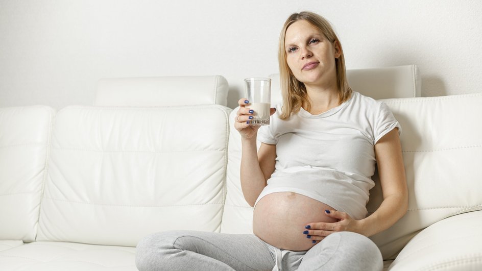 Беременная девушка сидит со стаканом на белом диване 