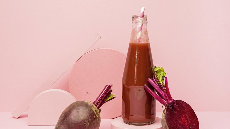 Бутылка свекольного кваса с трубочкой и два корнеплода свеклы на розовом фоне Бутылка свекольного кваса с трубочкой и два корнеплода свеклы на розовом фоне