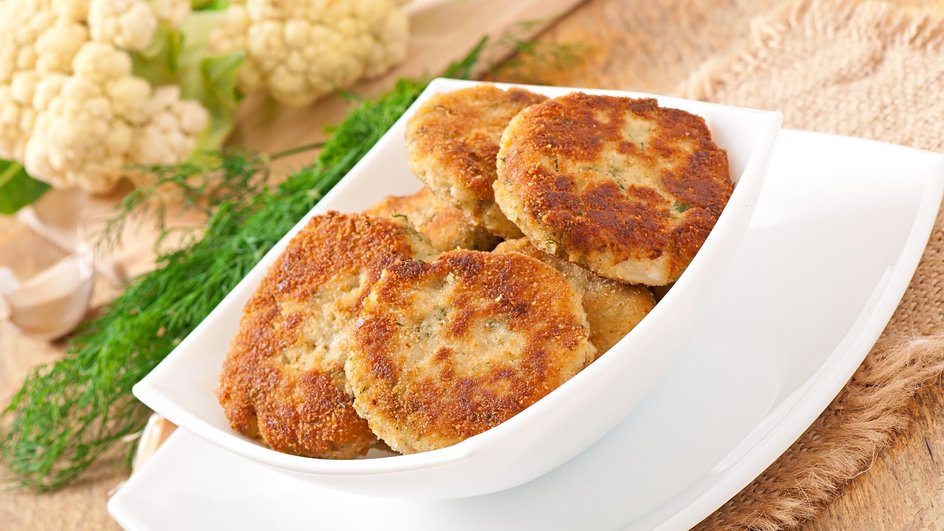 Биточки из кукурузной каши с цветной капустой лежат в белой керамической тарелке квадратной формы, которая стоит рядом с укропом и цветной капустой Биточки из кукурузной каши с цветной капустой лежат в белой керамической тарелке квадратной формы, которая стоит рядом с укропом и цветной капустой
