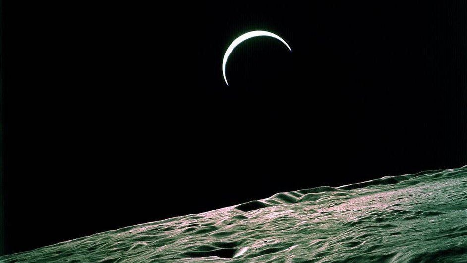 Земля появляется на черном небе в виде тонкого серпа бело-голубого цвета. На переднем плане — неровная каменистая поверхность Луны.