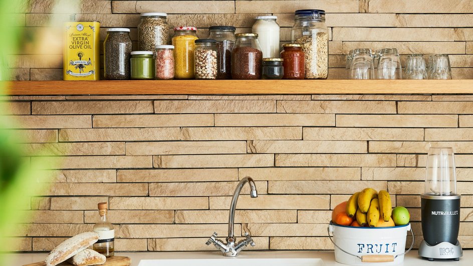 На стене на кухне открытая полка со специями, мойка, на столе стоит миска с фруктами