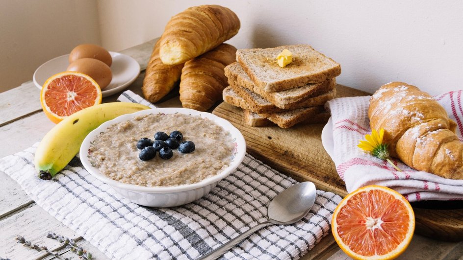 Тарелка овсяной каши с черникой на столе, рядом хлебцы, круассаны, яйца, банан и половинки апельсина.