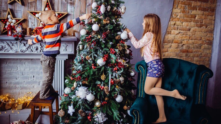 Мальчик и девочка украшают новогоднюю елку. Девочка стоит на синем диване, а мальчик - на табурете. За ними серый камин и деревянные звезды на стене с шариками внутри.