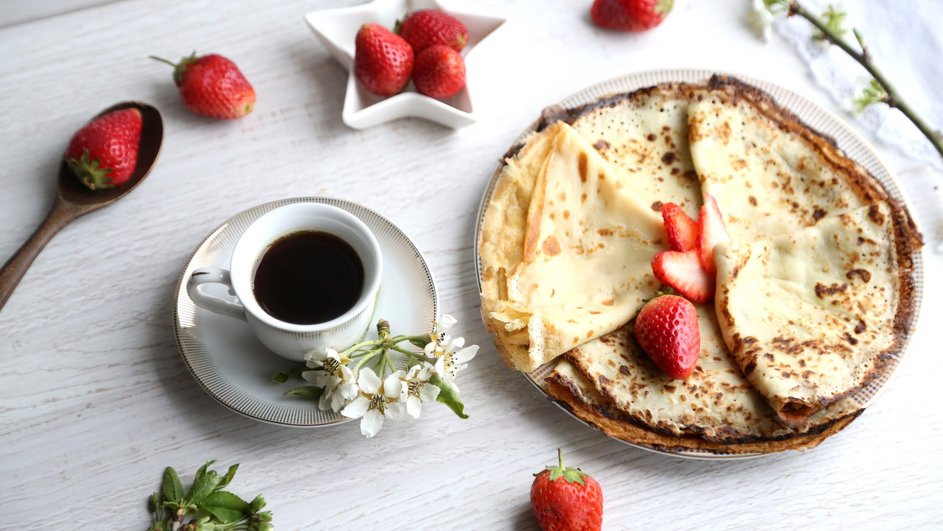 Вкусные блины без соды на кислом молоке лежат на тарелке рядом с чашкой чая и клубникой