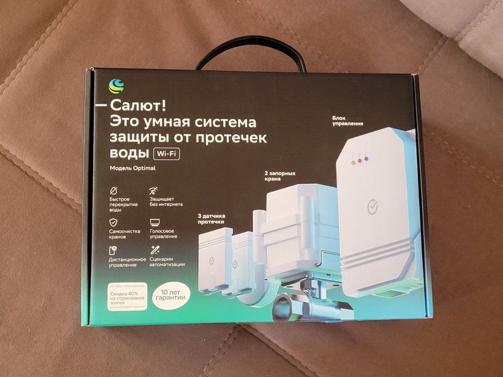 На коробке изображена умная система защиты от протечек воды с Wi-Fi. На передней части коробки написано: "Салют! Это умная система защиты от протечек воды. Модель Optimal". Также указаны функции: быстрое перекрытие воды, защита без интернета, самоочистка кранов, голосовое управление, дистанционное управление, сценарии автоматизации. В комплект входят 2 запорных крана и 3 датчика протечки. В правом нижнем углу указано, что предоставляется 10 лет гарантии. Также есть информация о скидке 40% на страхование жилья. На коробке изображена умная система защиты от протечек воды с Wi-Fi. На передней части коробки написано: "Салют! Это умная система защиты от протечек воды. Модель Optimal". Также указаны функции: быстрое перекрытие воды, защита без интернета, самоочистка кранов, голосовое управление, дистанционное управление, сценарии автоматизации. В комплект входят 2 запорных крана и 3 датчика протечки. В правом нижнем углу указано, что предоставляется 10 лет гарантии. Также есть информация о скидке 40% на страхование жилья.