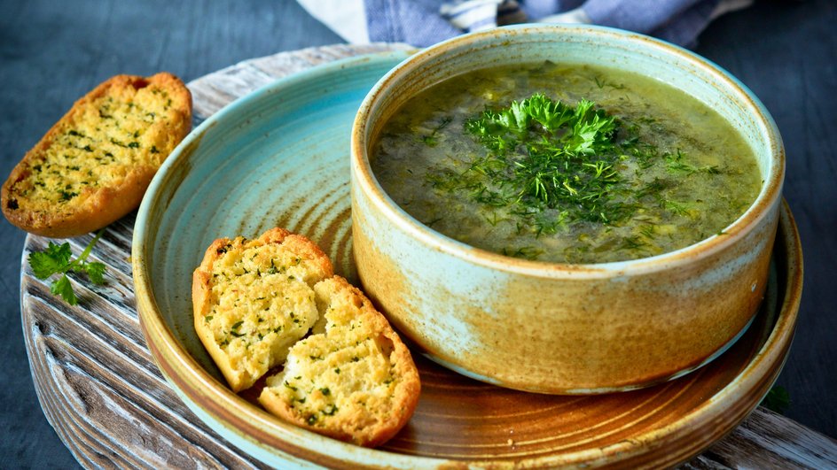 Щи с тушенкой и щавелем, украшенные зеленью, в супнице стоят на плоской тарелке рядом с кусочками хлеба на деревянной доске Щи с тушенкой и щавелем, украшенные зеленью, в супнице стоят на плоской тарелке рядом с кусочками хлеба на деревянной доске