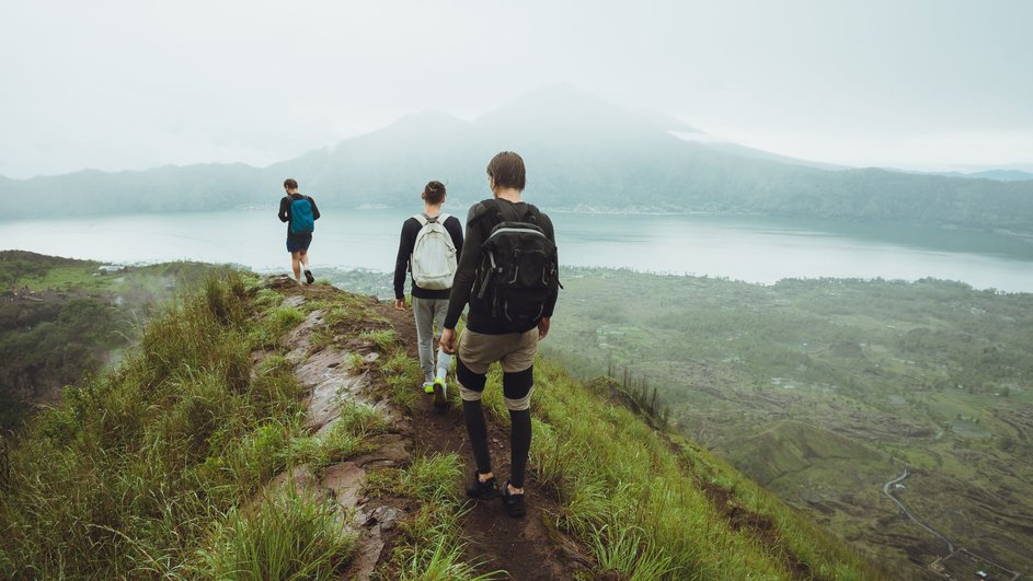 Три человека идут по холму с рюкзаками, на фоне белых облаков и вершины вулкана