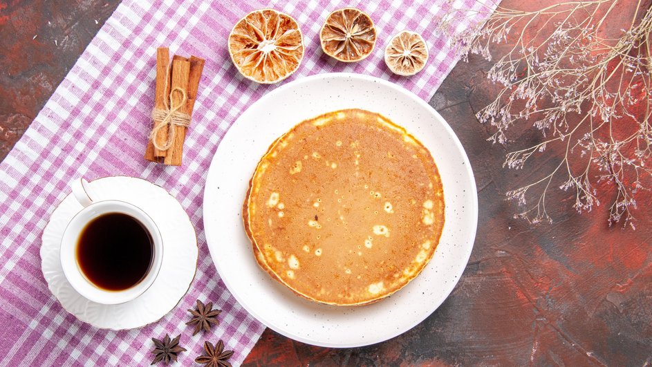 Заварные блины лежат на тарелке рядом с чашкой кофе, палочками корицы, сушенными апельсином и лимоном на столе с клетчатой скатертью