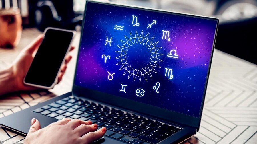 На экране ноутбука - знаки зодиака, рука держит телефон, а другая - на клавиатуре ноутбука.