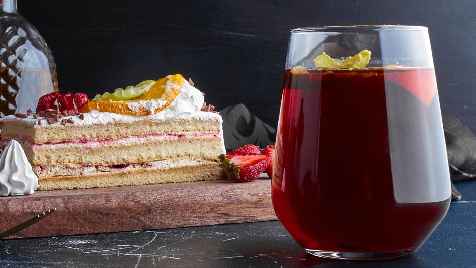 Стакан с компотом из сухофруктов, украшенным кусочками сухофруктов, рядом на деревянной доске лежит кусок торта Стакан с компотом из сухофруктов, украшенным кусочками сухофруктов, рядом на деревянной доске лежит кусок торта