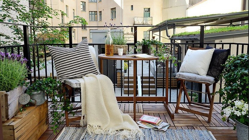 Открытый балкон с мебелью, подушками и растениями на фоне многоэтажного дома 