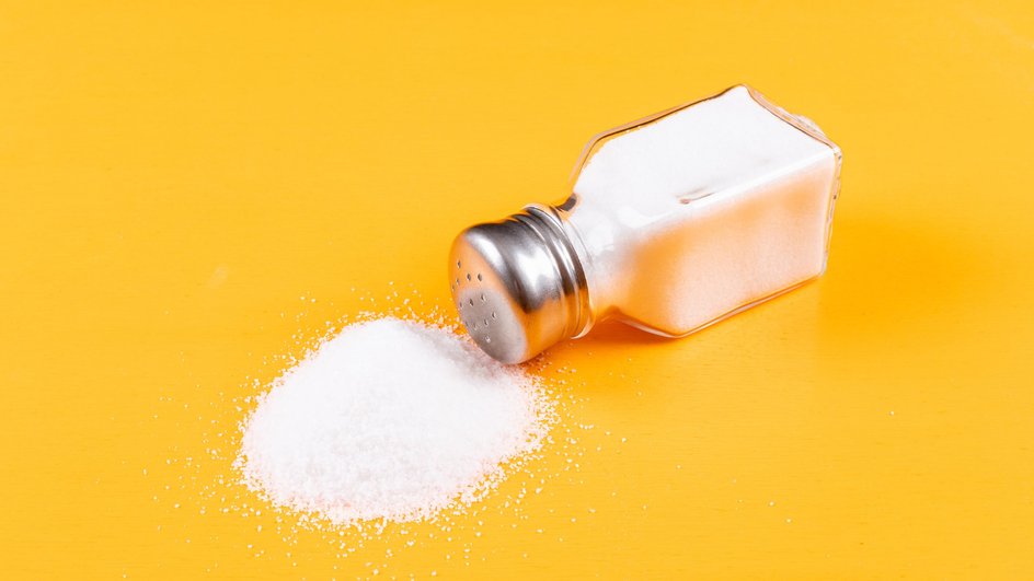 Прозрачная солонка с рассыпанной солью лежит на желтом столе 