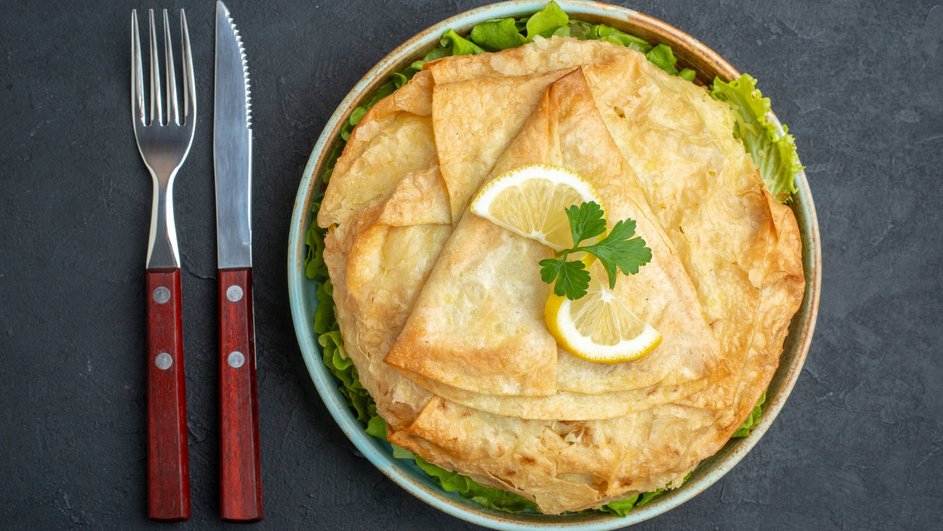 Кабачковые блины с сыром и чесноком, украшенные дольками лимона и зеленью, на тарелке лежат рядом с вилкой и столовым ножом с деревянной ручкой на темном столе Кабачковые блины с сыром и чесноком, украшенные дольками лимона и зеленью, на тарелке лежат рядом с вилкой и столовым ножом с деревянной ручкой на темном столе