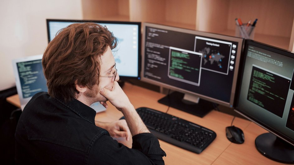 Молодой человек DevOps-инженер сидит за рабочим столом перед экранами компьютеров Молодой человек DevOps-инженер сидит за рабочим столом перед экранами компьютеров
