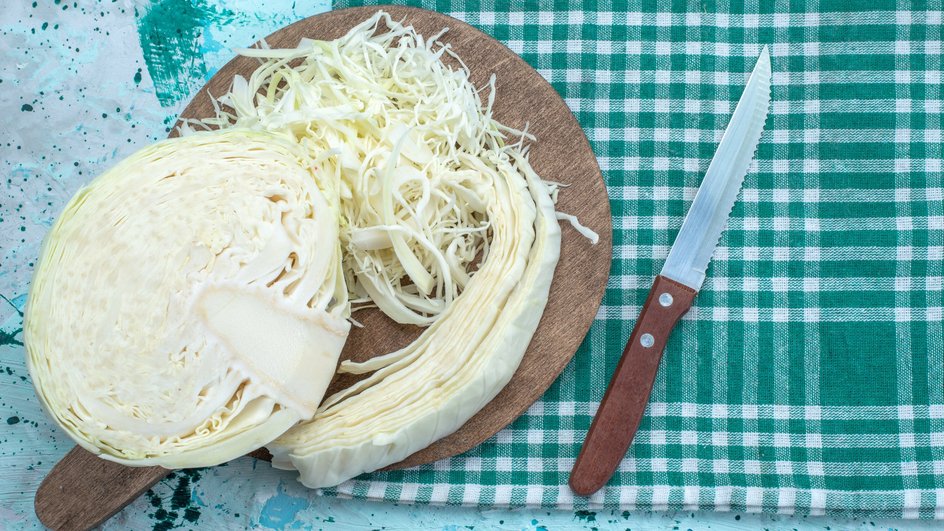 Капуста, нашинкованная тонкой соломкой, лежит на доске рядом с качаном капусты и ножом