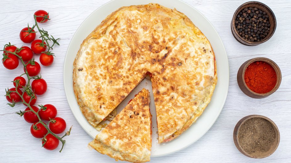 Лепешка с сыром, помидором и луком с лежащими рядом свежими овощами и приправами Лепешка с сыром, помидором и луком с лежащими рядом свежими овощами и приправами