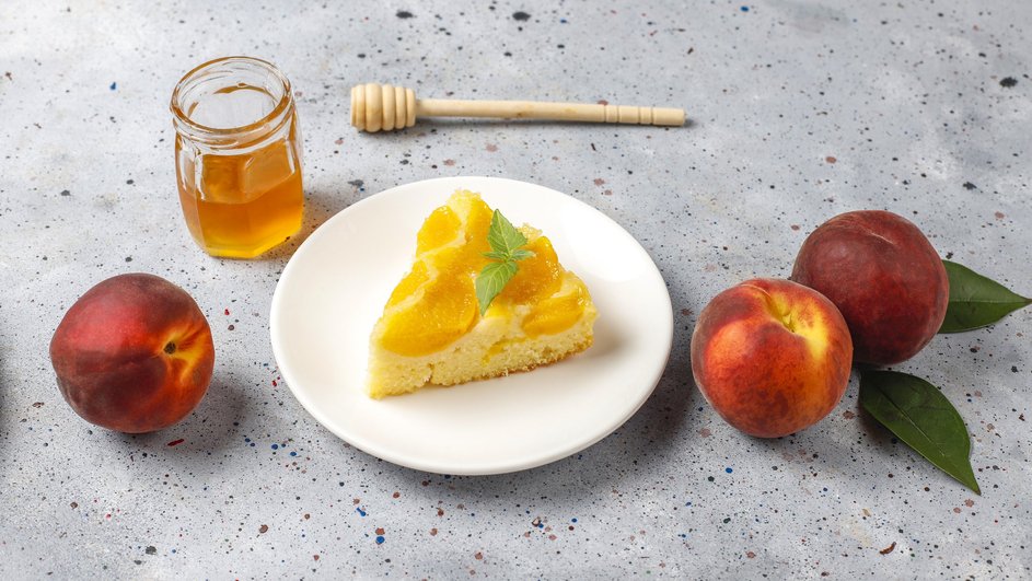 Кусочек шарлотки с персиками и корицей лежит на тарелке рядом с персиками и медом