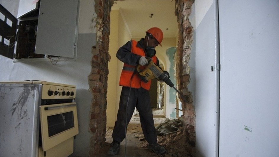 Рабочий в оранжевом жилете отбойным молотком долбит стену