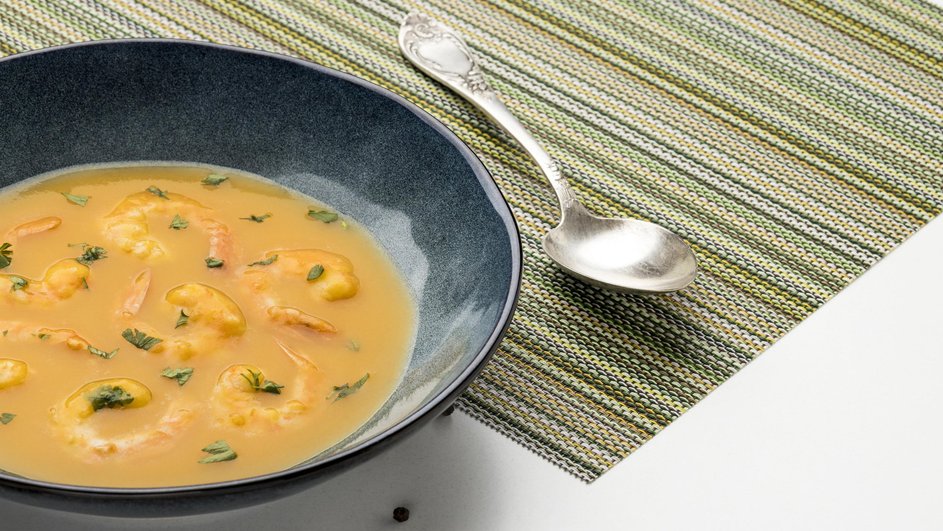 Вкусный суп с сыром, креветками и шампиньонами в тарелке стоит на столе рядом с ложкой и горошинами перца