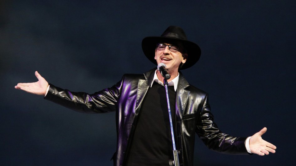 Михаил Боярский в шляпе и кожаном пиджаке разводит руки на сцене